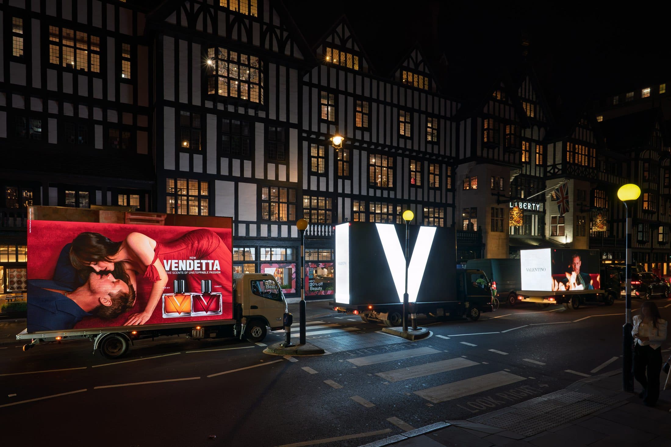 Three Monster Digital AdVans outside Liberty at night, the giant white V of the Valentino Vendetta campaign lit on the rear screen